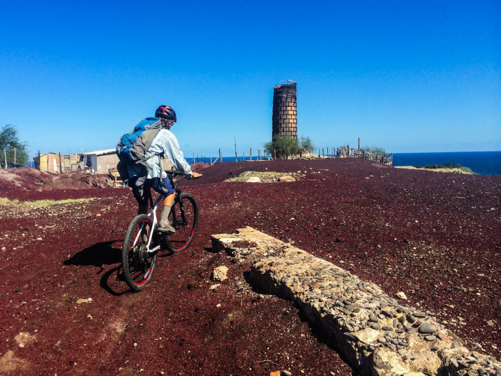 Josh rides out onto the flattened top of the tierra quemada cone, the red remnants of El Boleo Copper Company mine. The brick smokestack from the smelter stands at the edge of the pile.