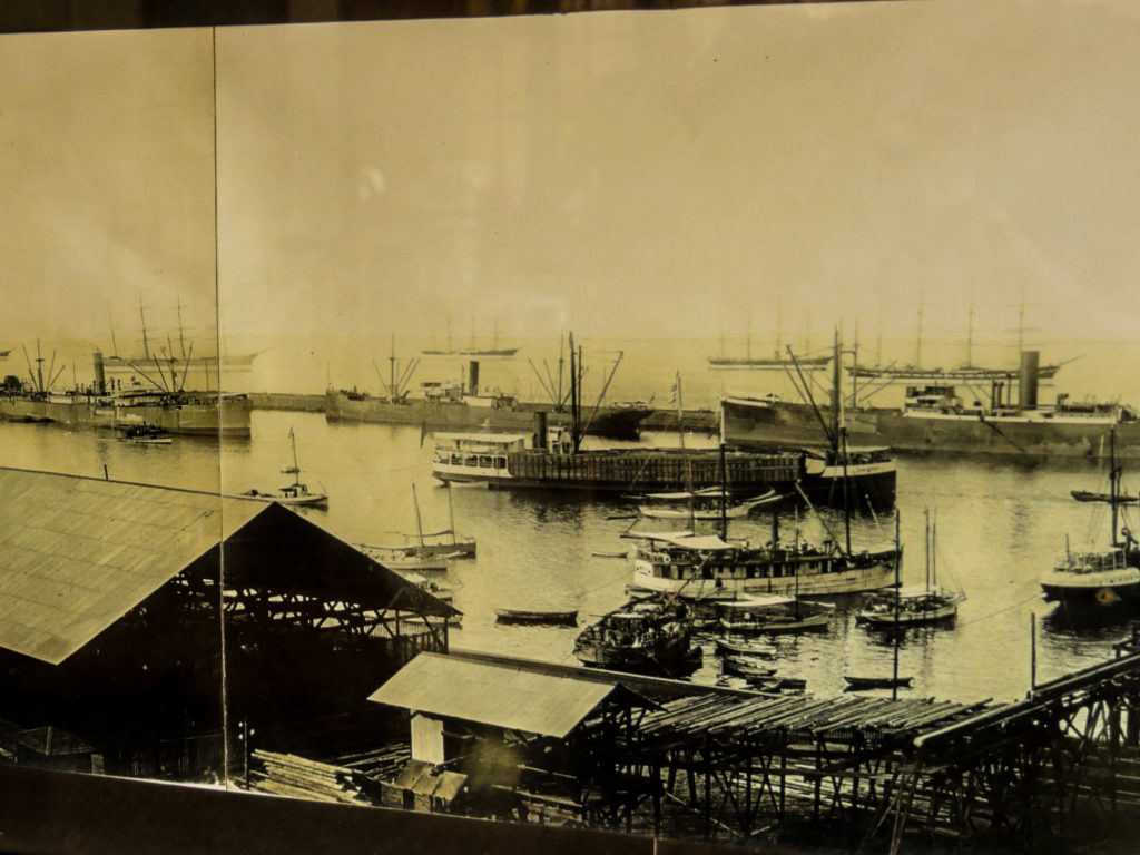 A photo of the harbor from the turn of the 20th century. Mexican-built steam ships designed to cross the Sea of Cortez sit in the harbor, and international sailing ships sit outside the breakwater wall, waiting to offload giant wooden beams and take on the mined copper.