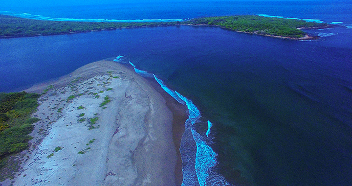 The narrow estuary entrance to Aserradores. The channel, marked by the riffles exiting the estuary near the top right of the photo, is flanked by rocks to starboard and breaking waves to port.